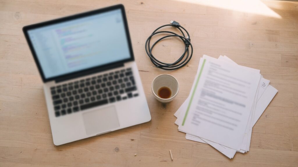 A laptop and a short stack of printed pages on a desk, the screen too soft to read